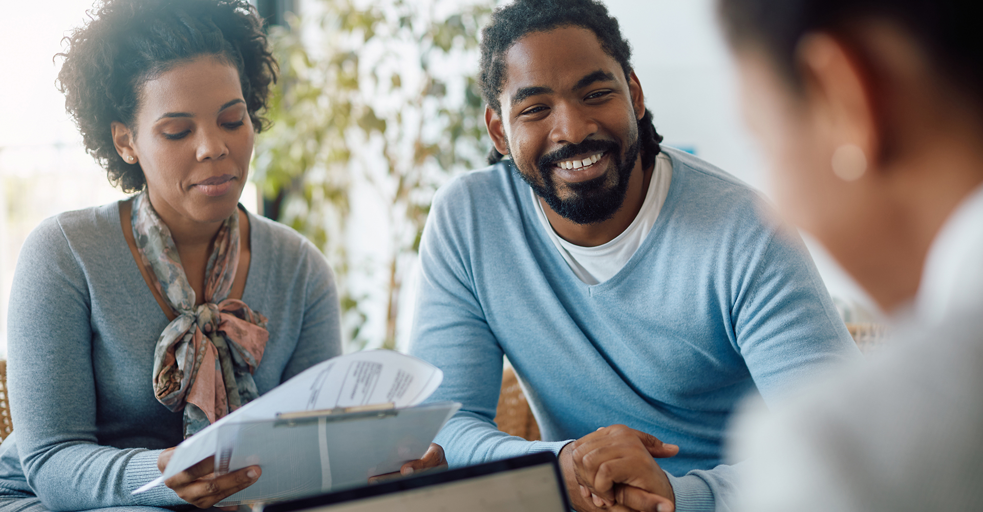 A smiling couple sits across from a professional during a meeting. The woman is holding a clipboard with documents, while the man listens attentively with a cheerful expression.