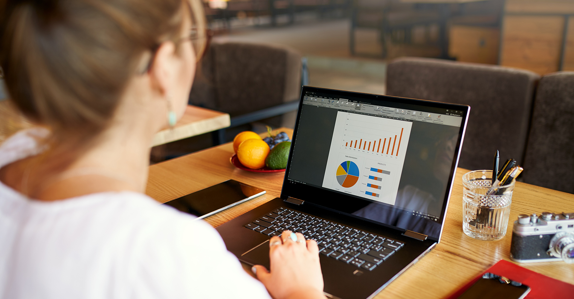 A woman works at a wooden table on a laptop displaying colorful charts and graphs, including a bar graph, pie chart, and stacked bar charts. The setting appears to be a casual workspace or café,.