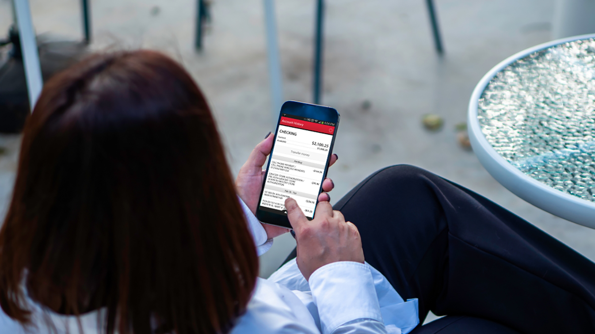 An individual holds a smartphone and uses a mobile banking app; the screen displays account savings and transaction options.