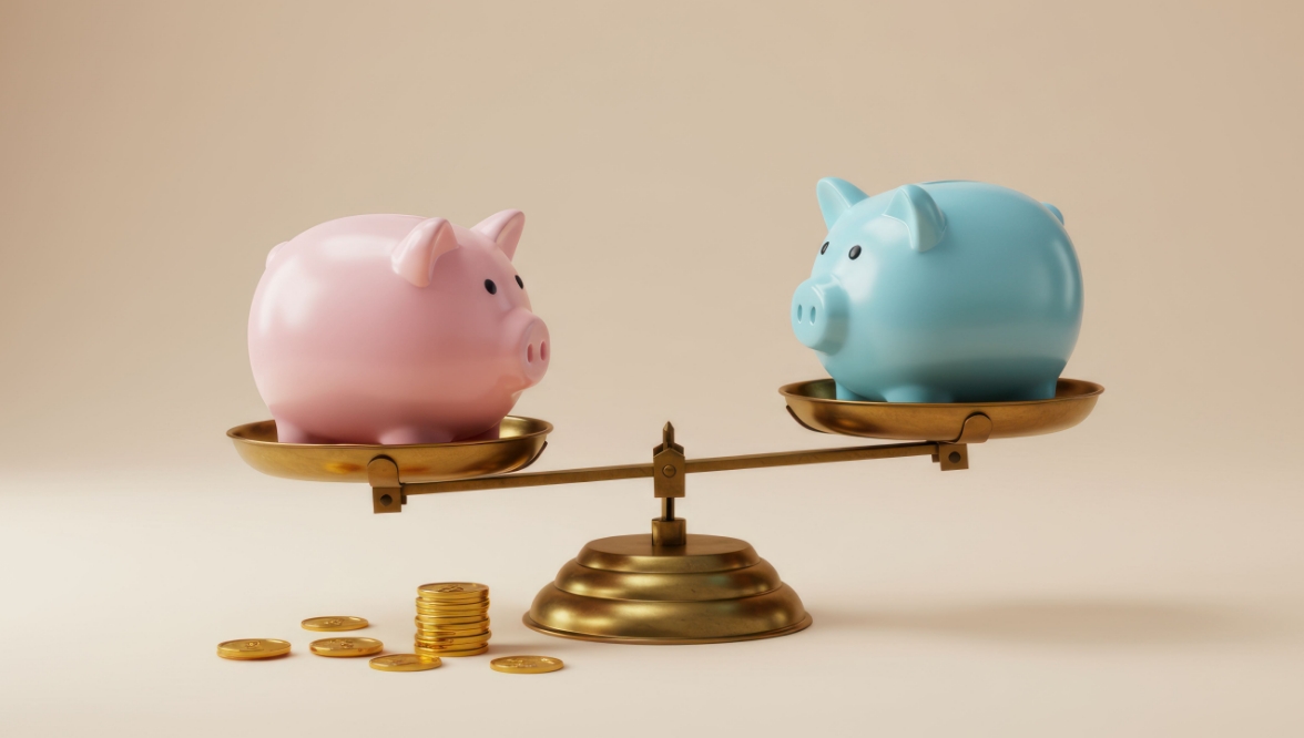 A pink piggy bank and a blue piggy bank sit on either side of a brass balance, with gold coins near the pink bank.