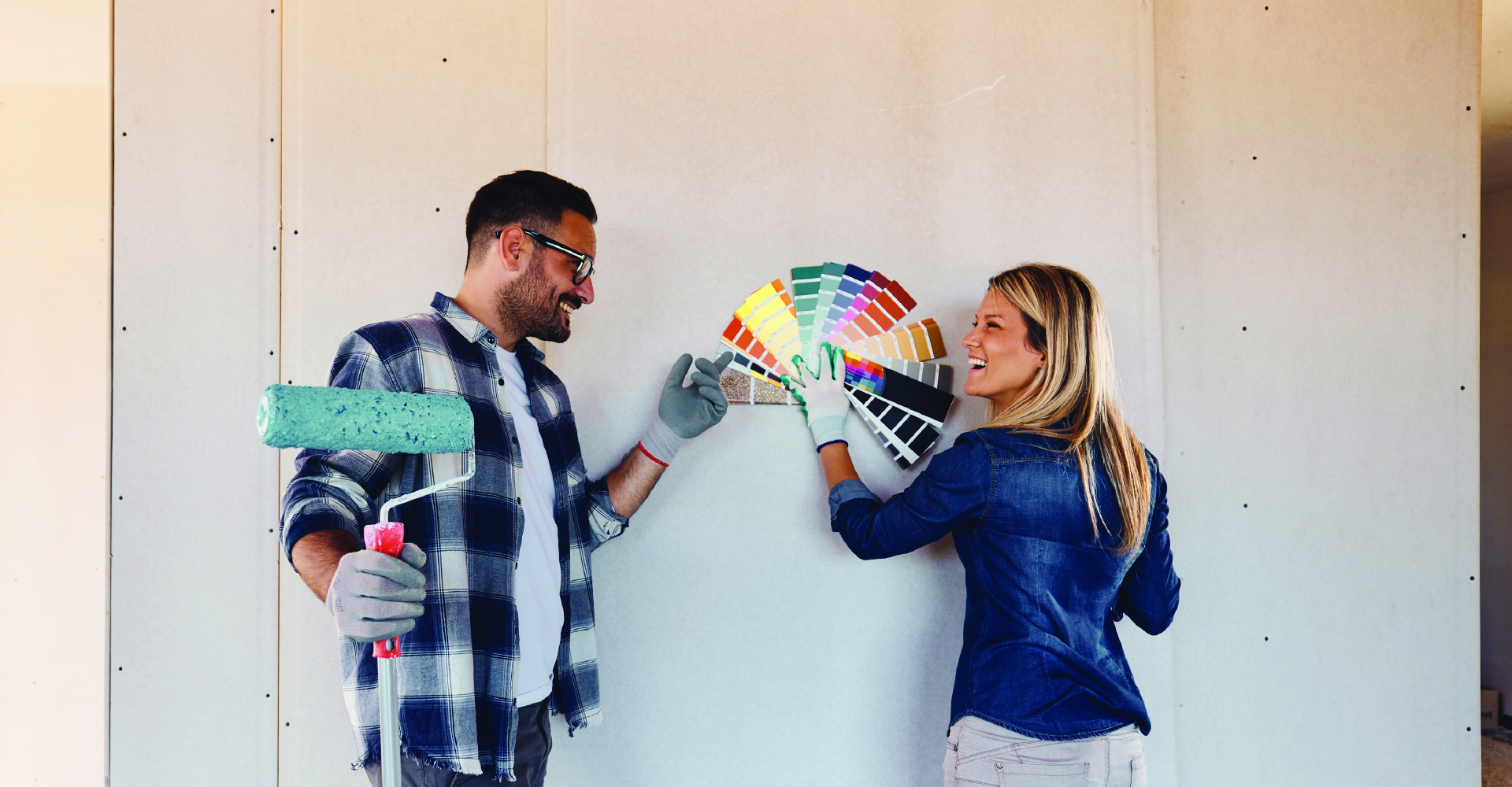 A young couple is standing in a partially renovated room, discussing paint colors while one person holds a fan of color swatches.