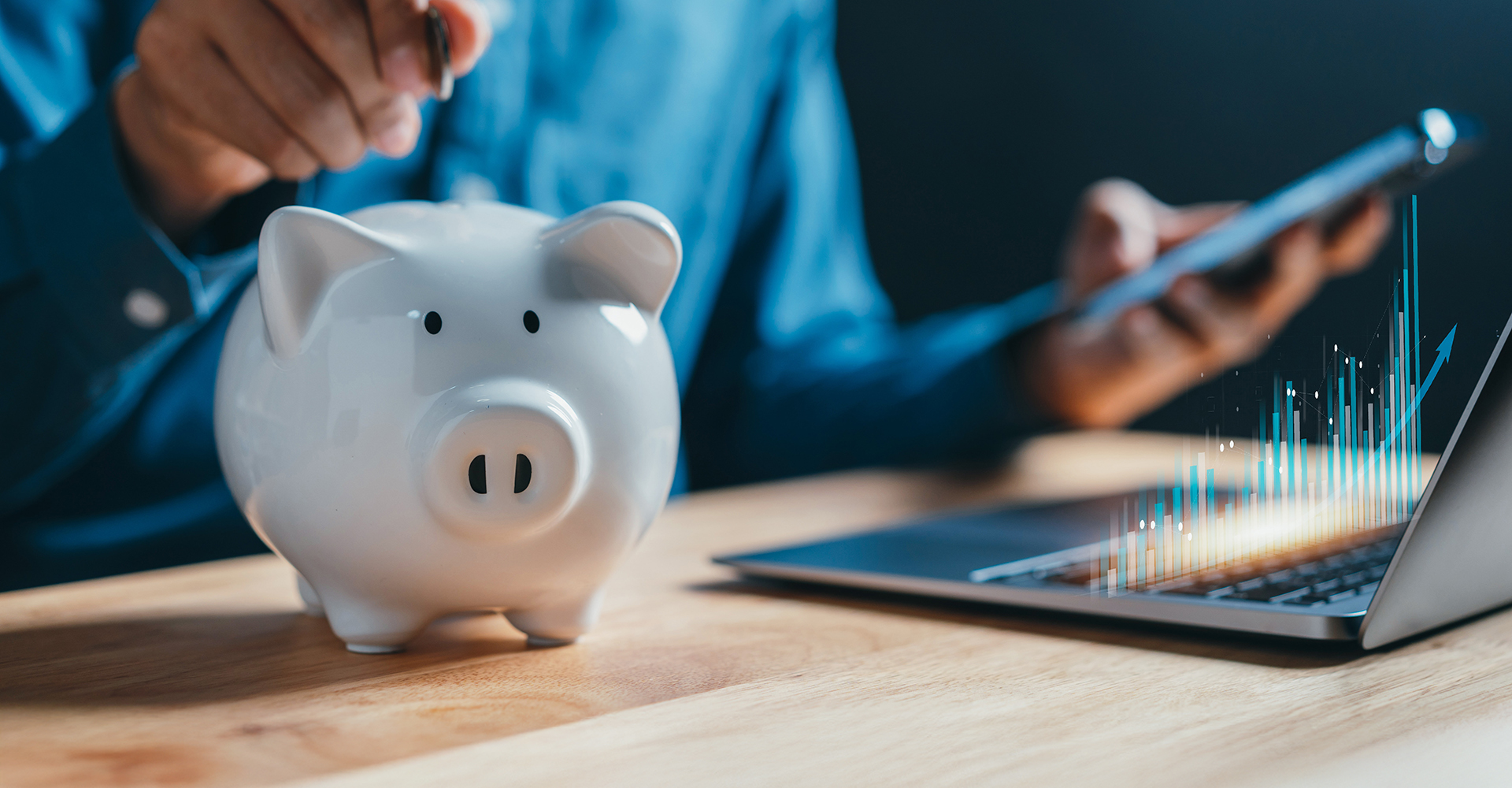 A man in a blue button-up shirt drops a coin into a porcelain piggy bank while looking at his cell phone, an open laptop showing graphs on the table in front of him.