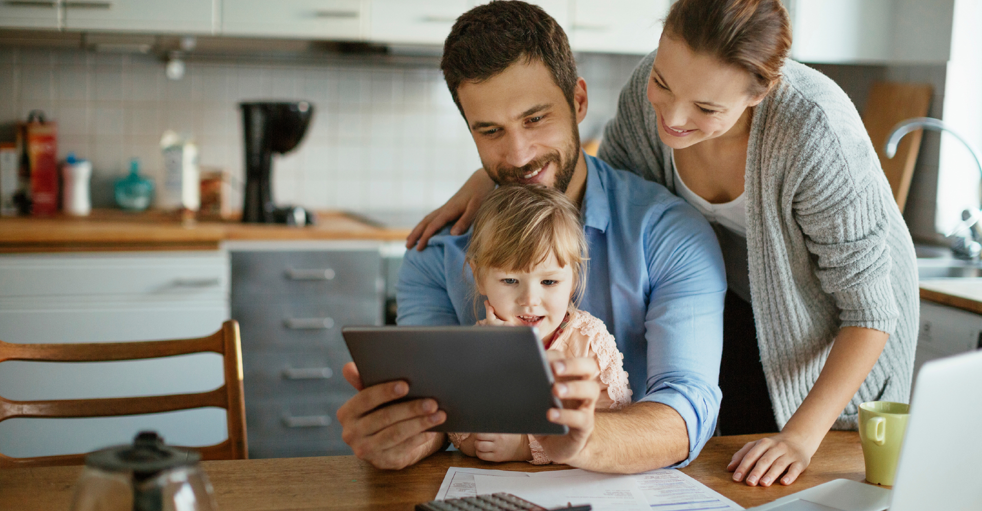 A young woman and a young man with a child on his lap look at a tablet and smile in a kitchen setting. An open laptop, papers, a calculator, and a coffeepot are on the table in front of them.