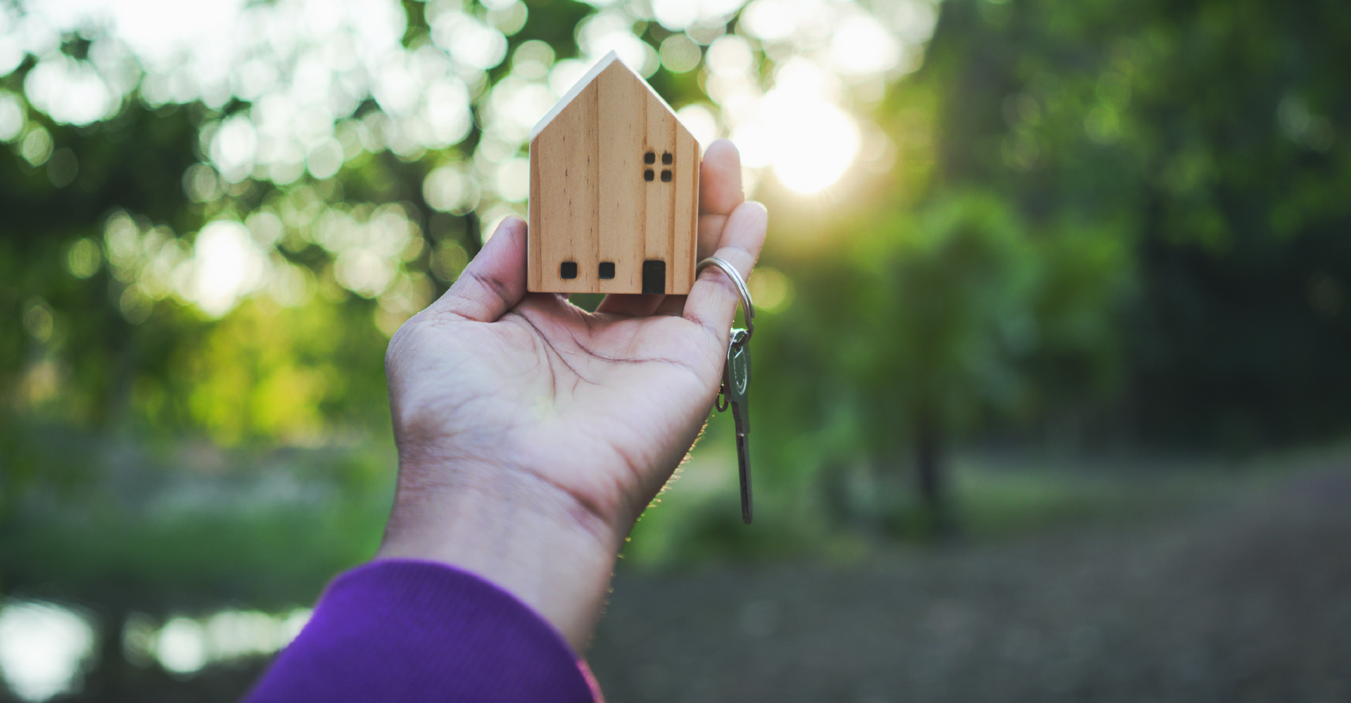A man's hand holding a small wooden model house and key up towards the sun.